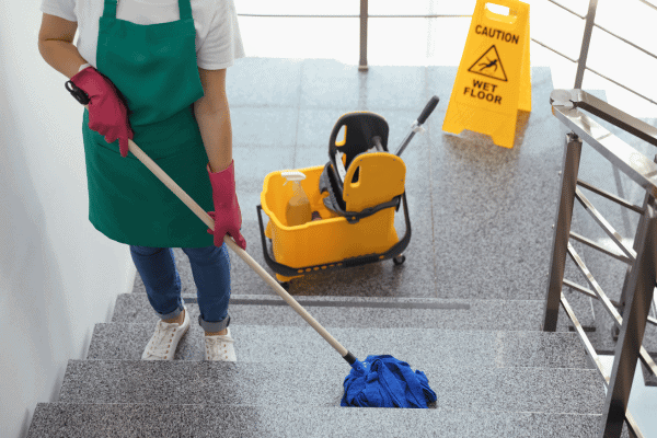 A woman mopping stairs with a caution wet floor sign and a mop bucket nearby.