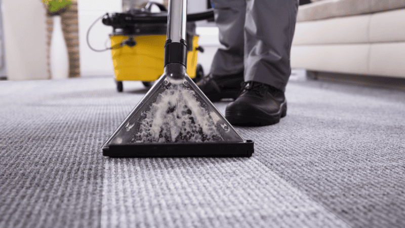 A professional carpet cleaner uses a steam cleaning machine to deep clean a gray carpet in a modern living room.