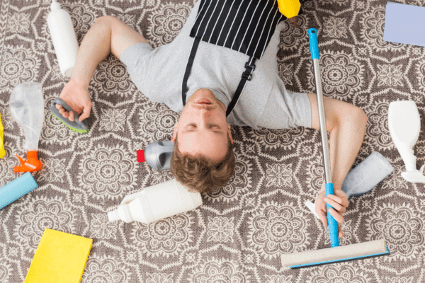 A tired man in an apron lies on a patterned carpet surrounded by cleaning supplies after a cleaning session.
