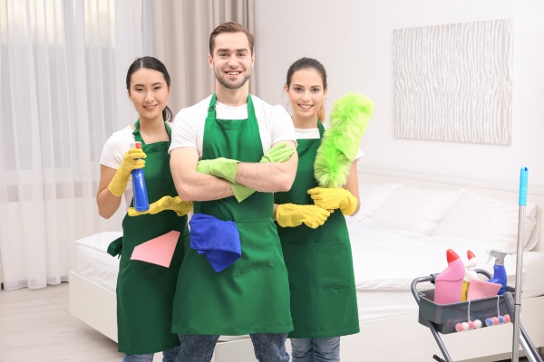 Three cleaning professionals in aprons with cleaning supplies and cart.