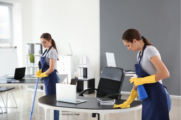 Two women cleaning office desks with laptops.